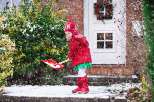 Little girl shoveling snow on home drive way. Beautiful house decorated for Christmas. Child with shovel playing outdoors in Xmas season. Family removing snow during blizzard. Kids play outside.
